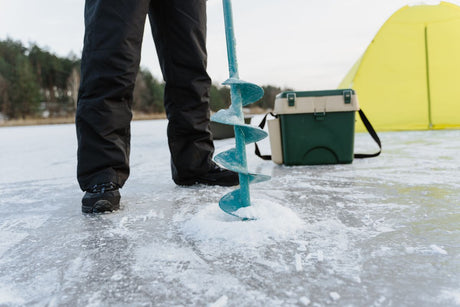 Hand auger cutting a fresh hole in bare lake ice beside a cooler and shelter